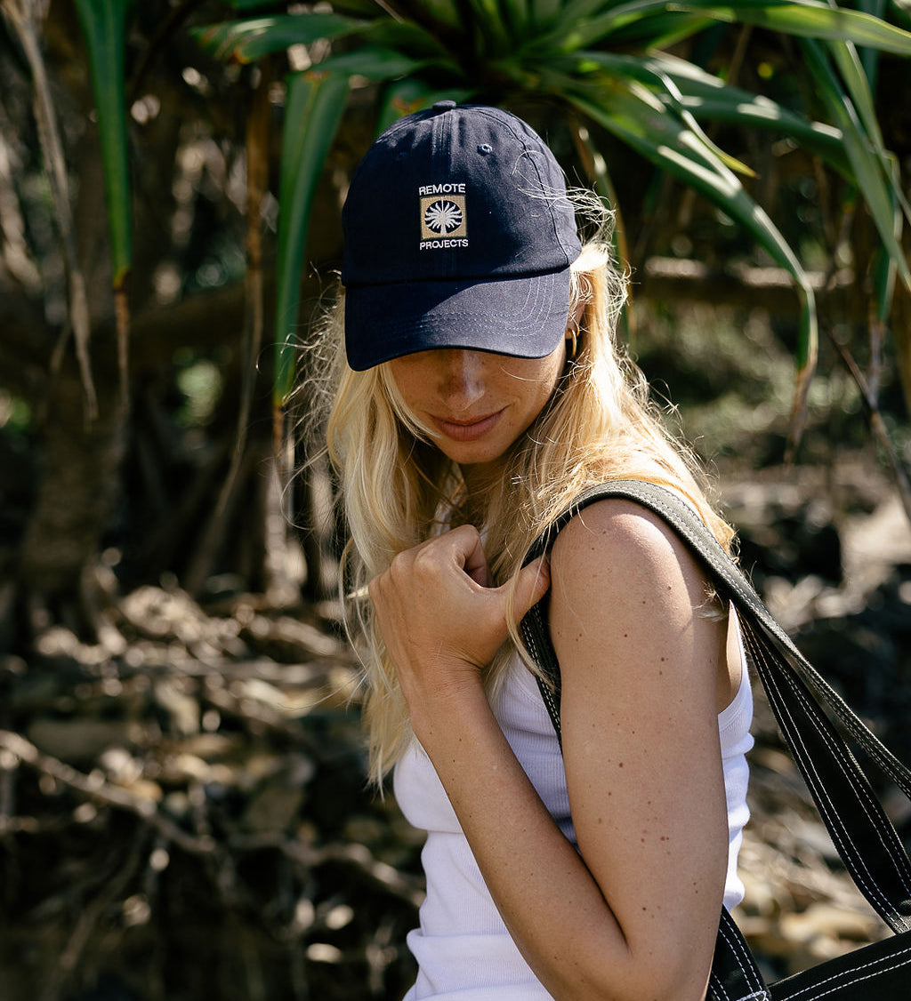 A woman outdoors wearing a navy blue baseball cap with a grass tree logo, a white tank top, and blue jeans, carrying a black tote bag.