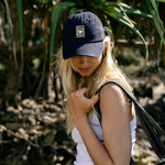 A woman outdoors wearing a navy blue baseball cap with a grass tree logo, a white tank top, and blue jeans, carrying a black tote bag.