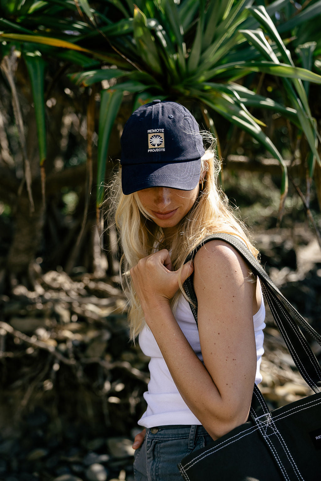 A woman outdoors wearing a navy blue baseball cap with a grass tree logo, a white tank top, and blue jeans, carrying a black tote bag.