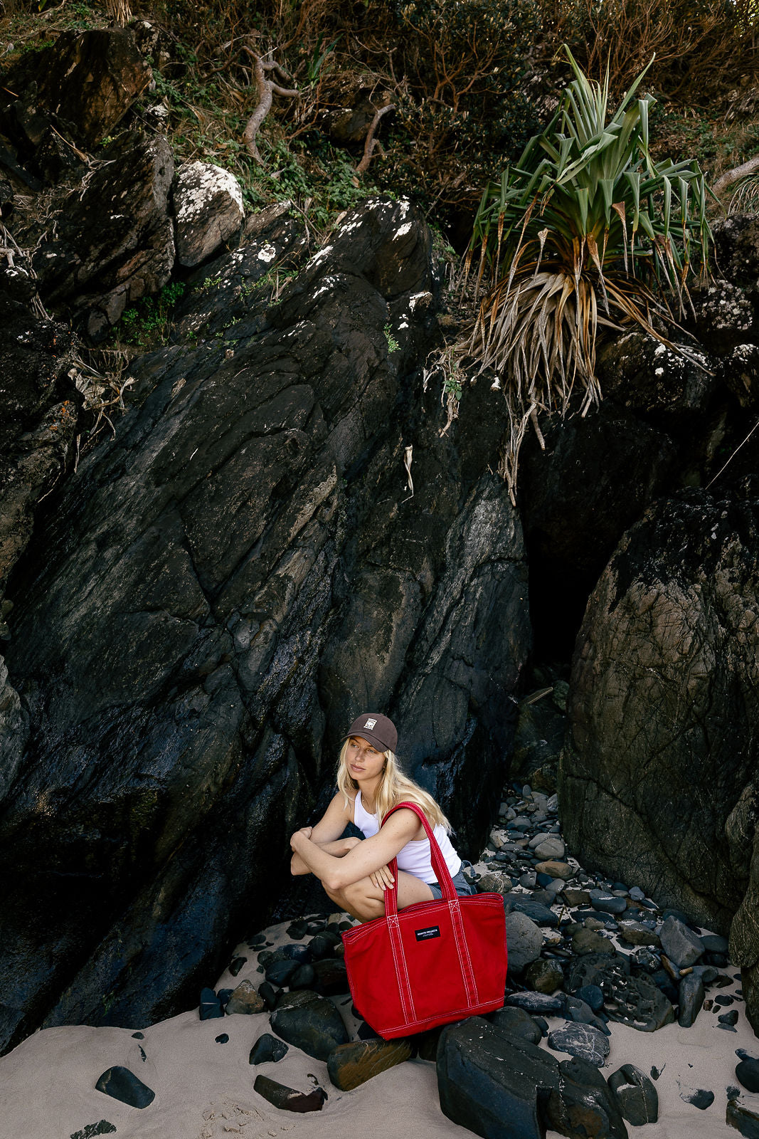 A woman sits on a rocky beach next to a large red tote bag, with dark cliffs and green plants in the background.