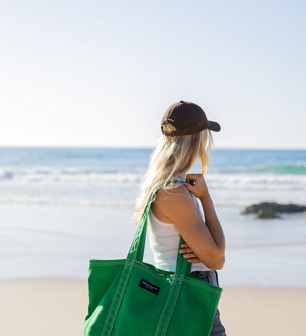A woman on a beach carries a large green canvas tote bag over her shoulder. She is seen from behind, wearing a brown cap, a white tank top, and denim shorts, with the ocean and sandy beach in the background.