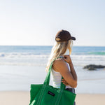 A woman on a beach carries a large green canvas tote bag over her shoulder. She is seen from behind, wearing a brown cap, a white tank top, and denim shorts, with the ocean and sandy beach in the background.
