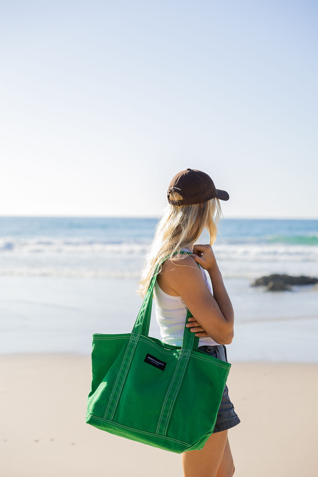 A woman on a beach carries a large green canvas tote bag over her shoulder. She is seen from behind, wearing a brown cap, a white tank top, and denim shorts, with the ocean and sandy beach in the background.